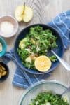 A blue bowl sitting on a blue striped towel with kale salad inside and a half lemon cut side up.