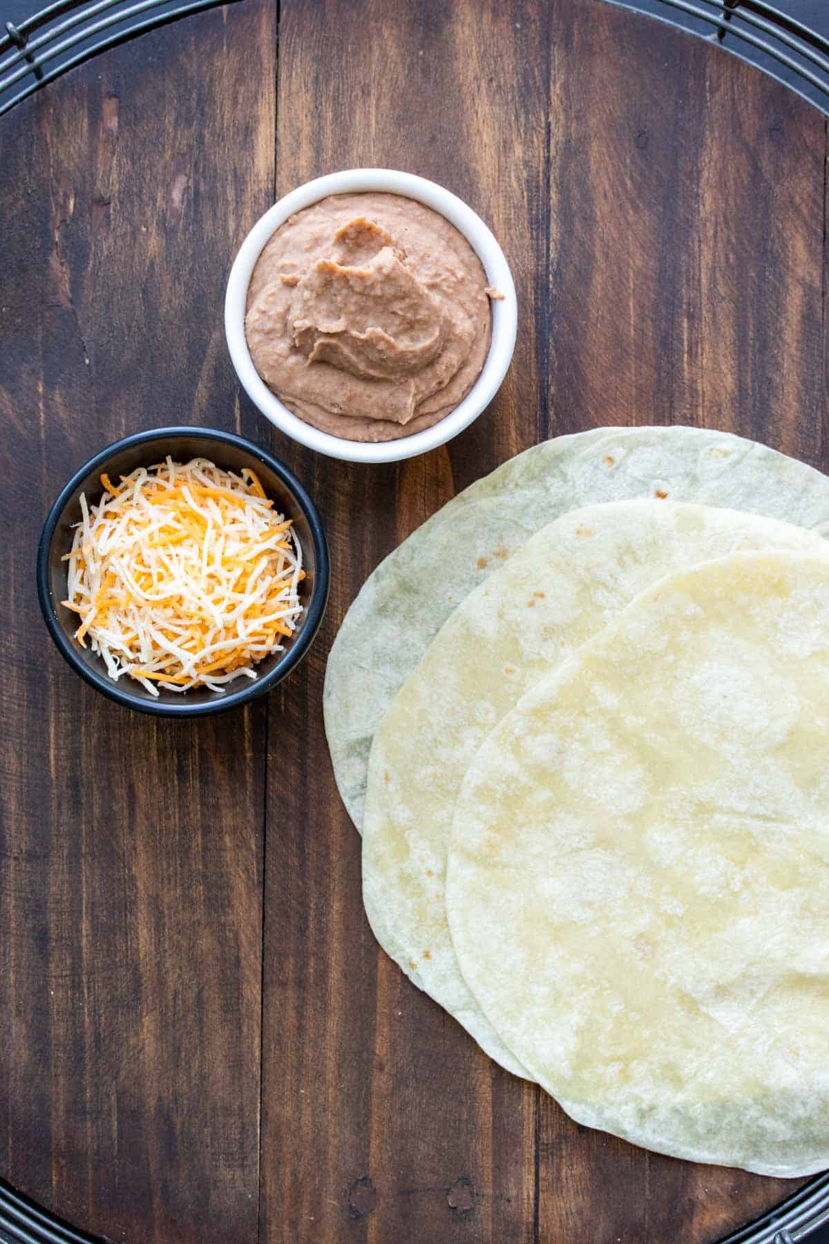 Photo of tortillas and bowls of refried beans and shredded cheese on a wooden surface