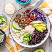 A cream colored bowl with a taco bowl inside on a wooden surface surrounded by other ingredients