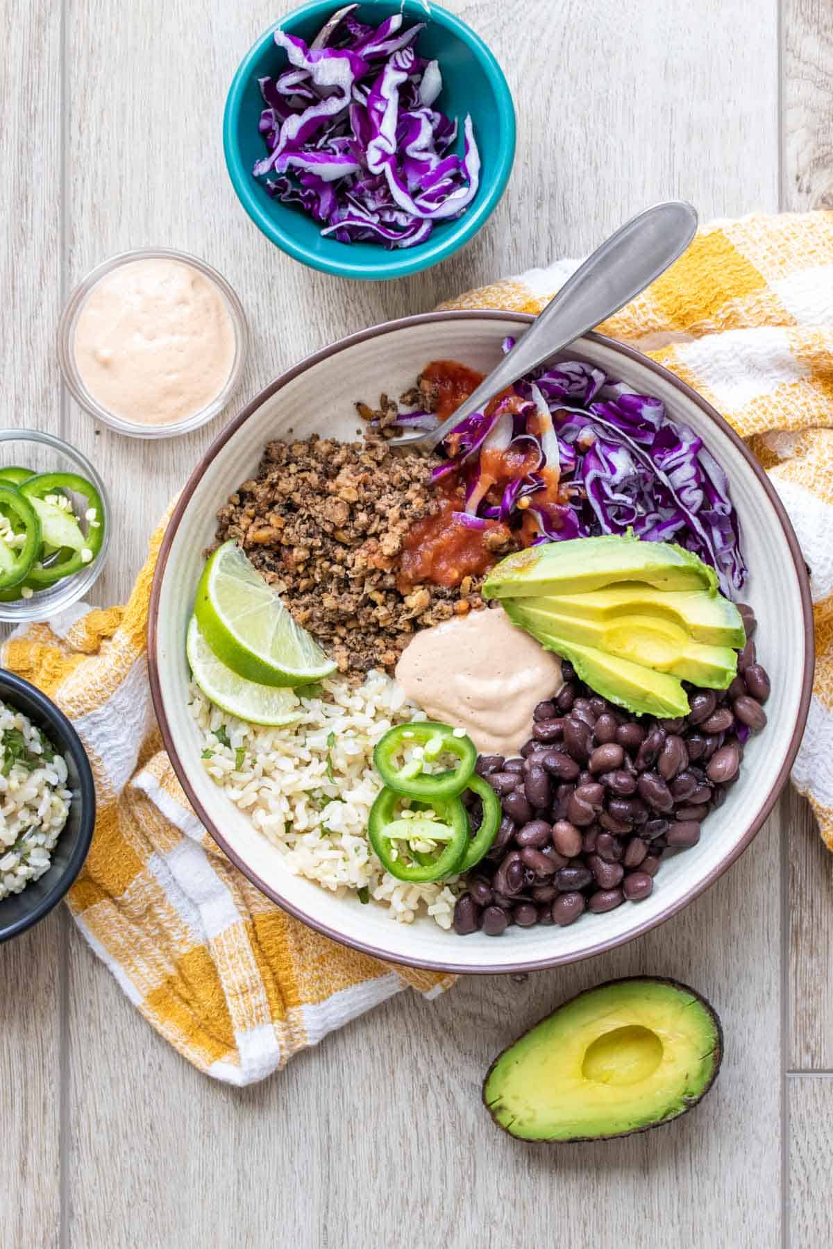A cream colored bowl with a taco bowl inside on a wooden surface surrounded by other ingredients