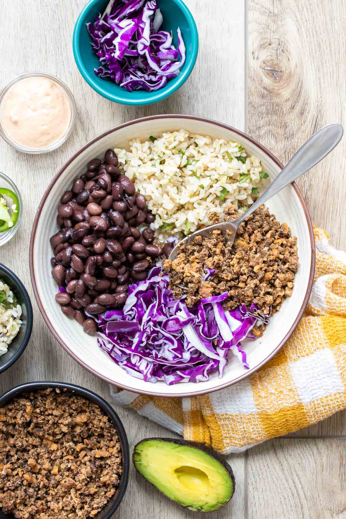 A cream colored bowl with rice, lentil taco meat, beans and cabbage inside on a light wooden surface