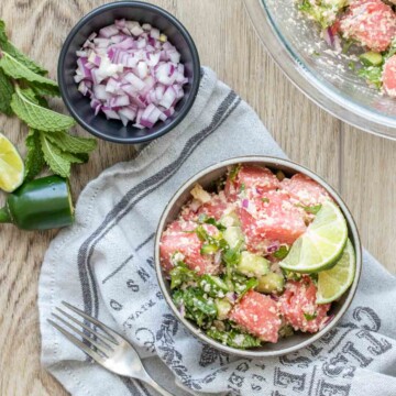 A bowl of watermelon feta mint salad next to a glass bowl of it and slices of onion, jalapeno and lime