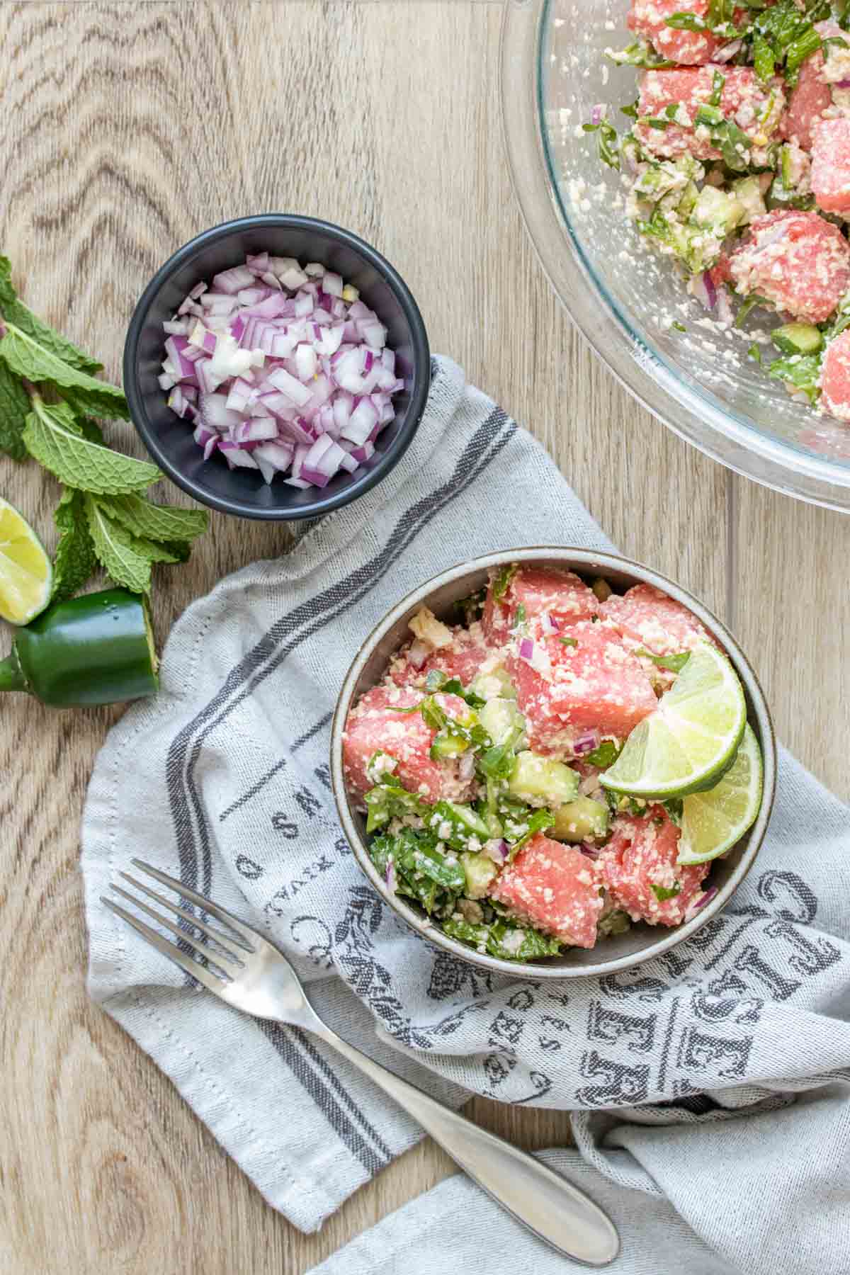 A bowl of watermelon feta mint salad next to a glass bowl of it and slices of onion, jalapeno and lime.