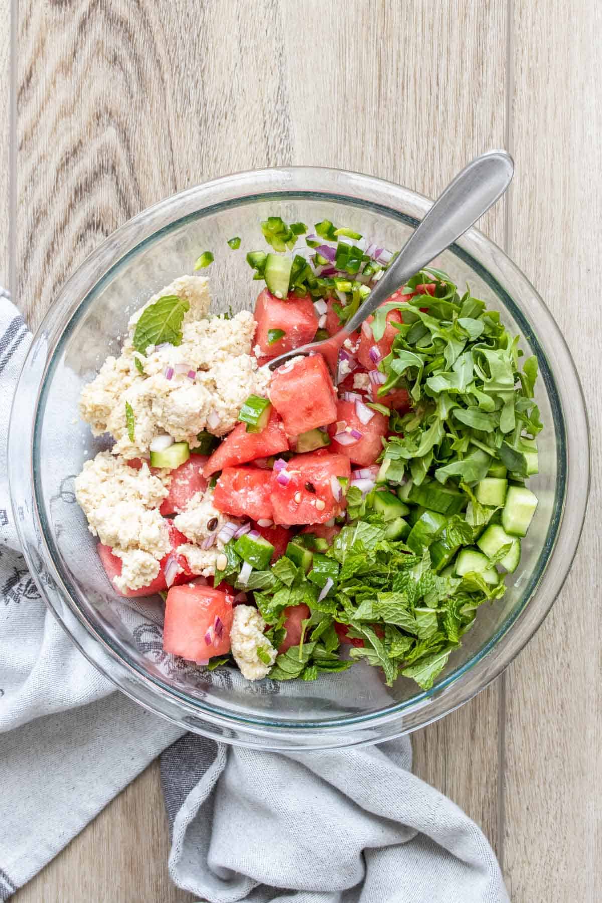 A glass bowl with watermelon, feta, cucumber, arugula and mint.