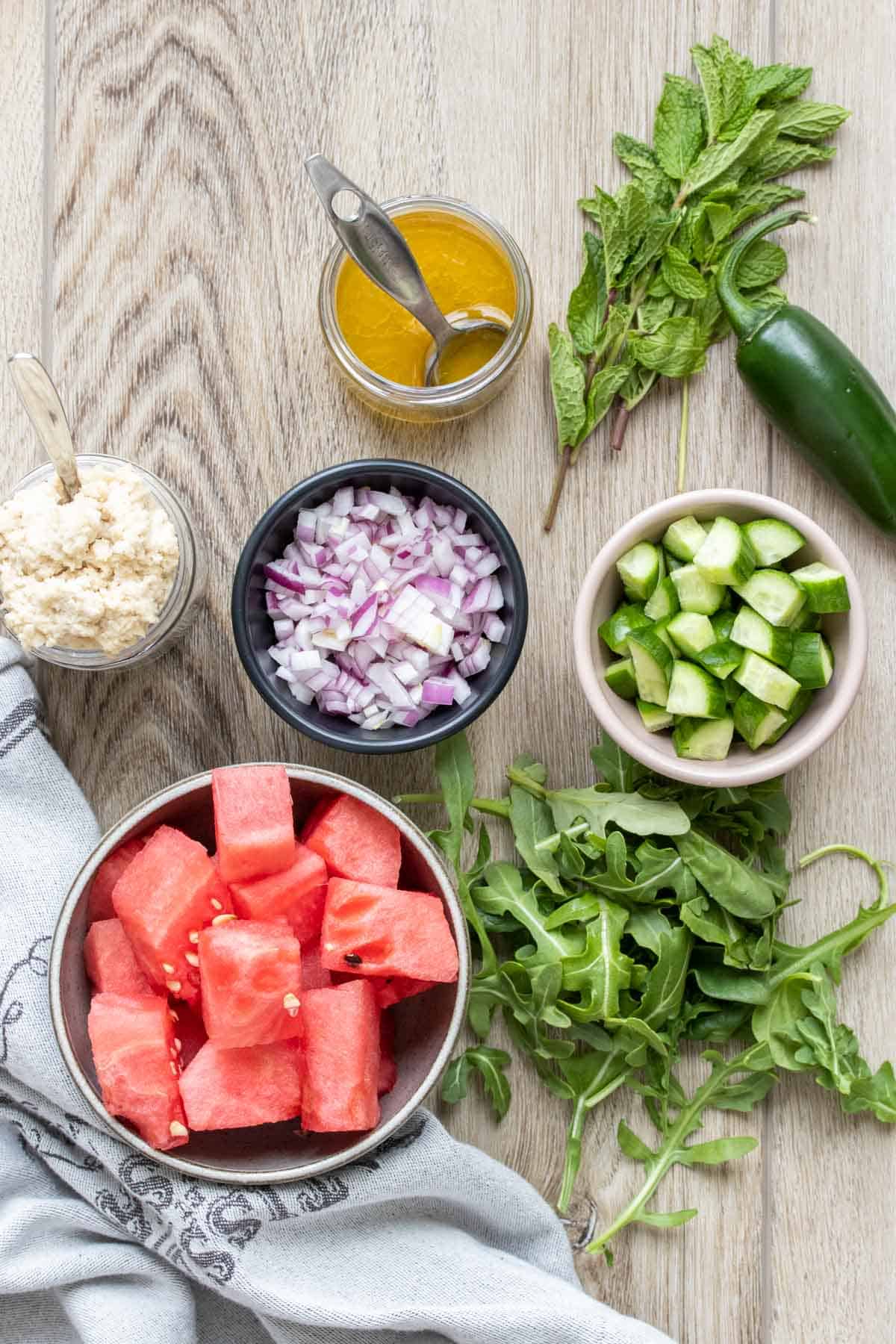 Bowls with ingredients to make a watermelon feta mint salad on a wooden surface.