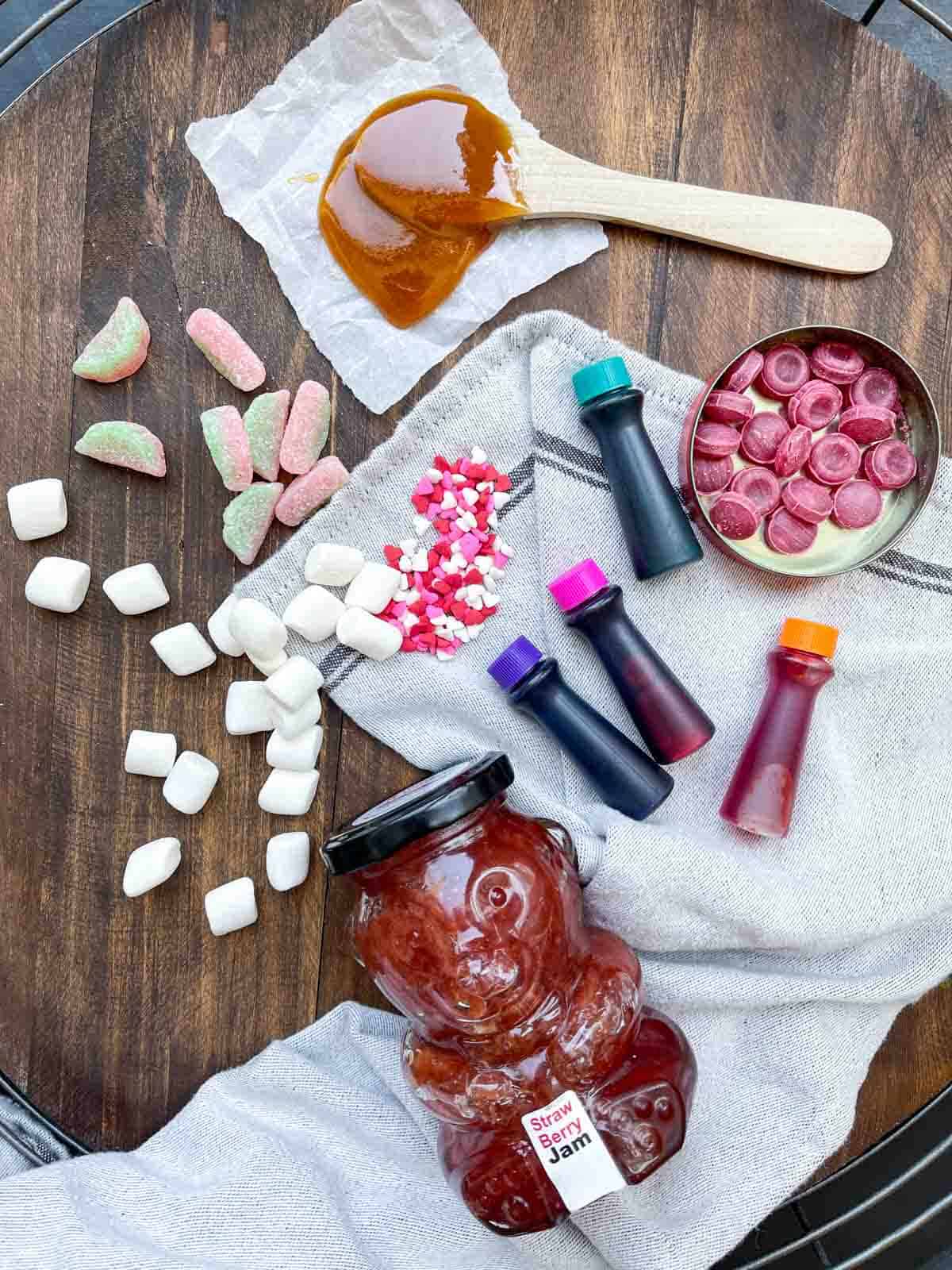 Top view of a wooden surface with food coloring, candy, a spoonful of honey and a bear jam jar