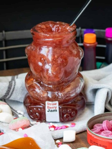 A bear shaped glass jar filled with strawberry jam surrounded by other ingredients