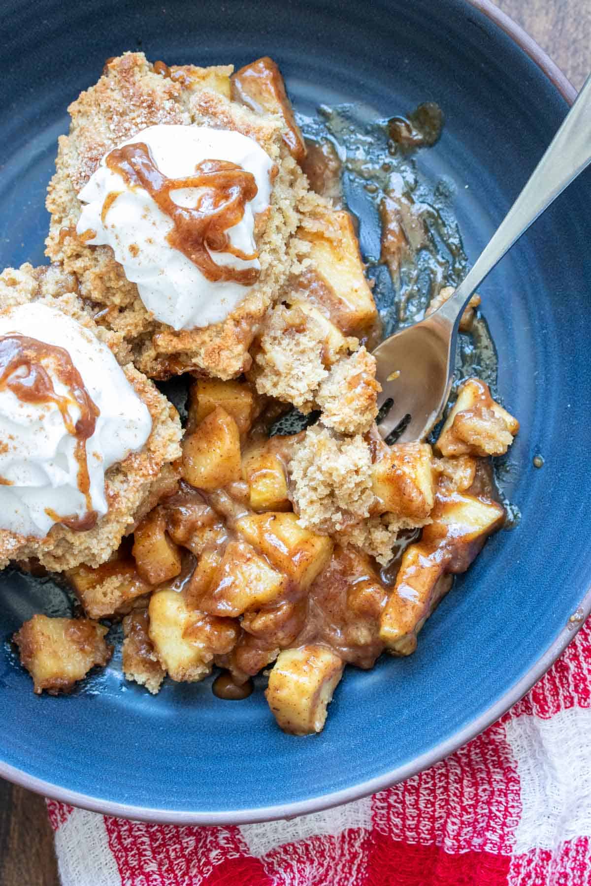 Fork eating apple cobbler with whipped cream in a dark blue bowl