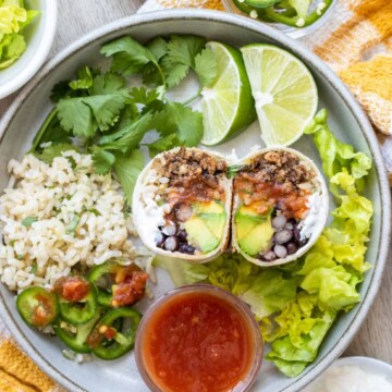 A burrito that has been cut in half sitting on a grey plate next to ingredients