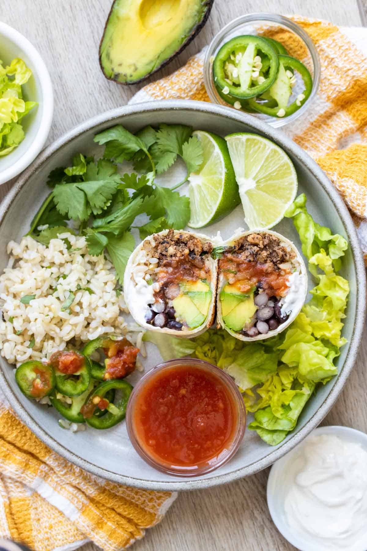 A burrito that has been cut in half sitting on a grey plate next to ingredients