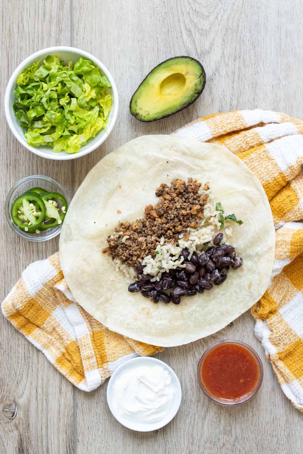 A tortilla on a yellow checked towel with rice, beans and veggie taco meat in the center of it