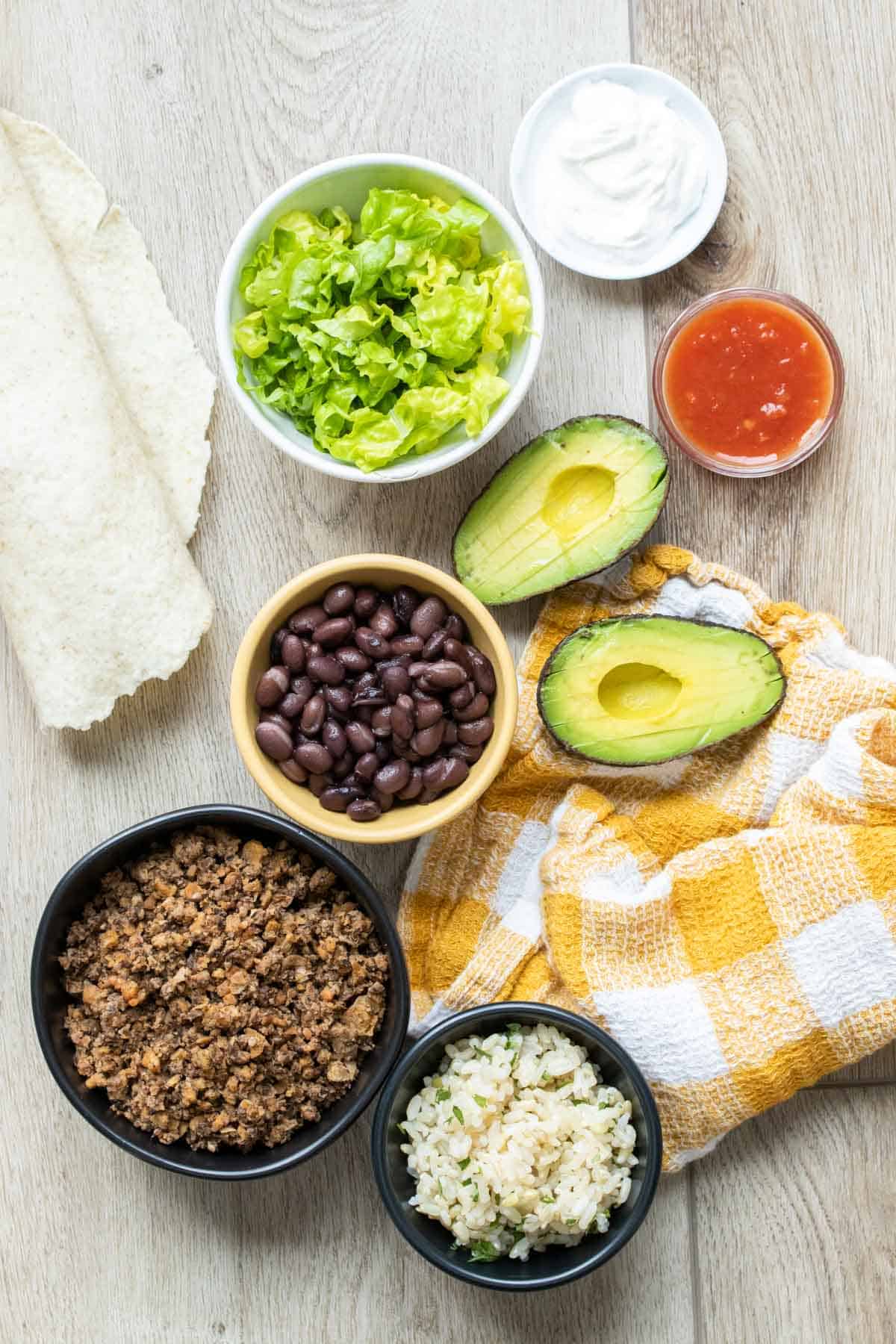 Ingredients for a burrito laying on a wooden surface with a yellow checked towel