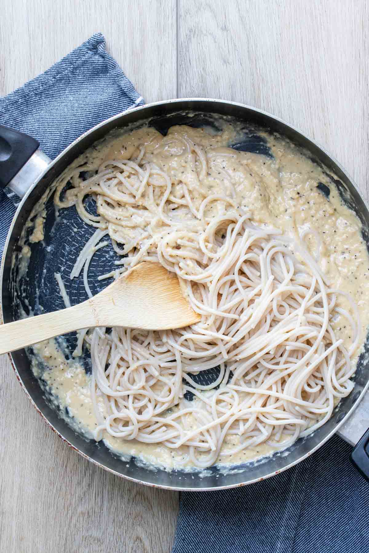Wooden spoon mixing spaghetti into a creamy sauce in a saute pan