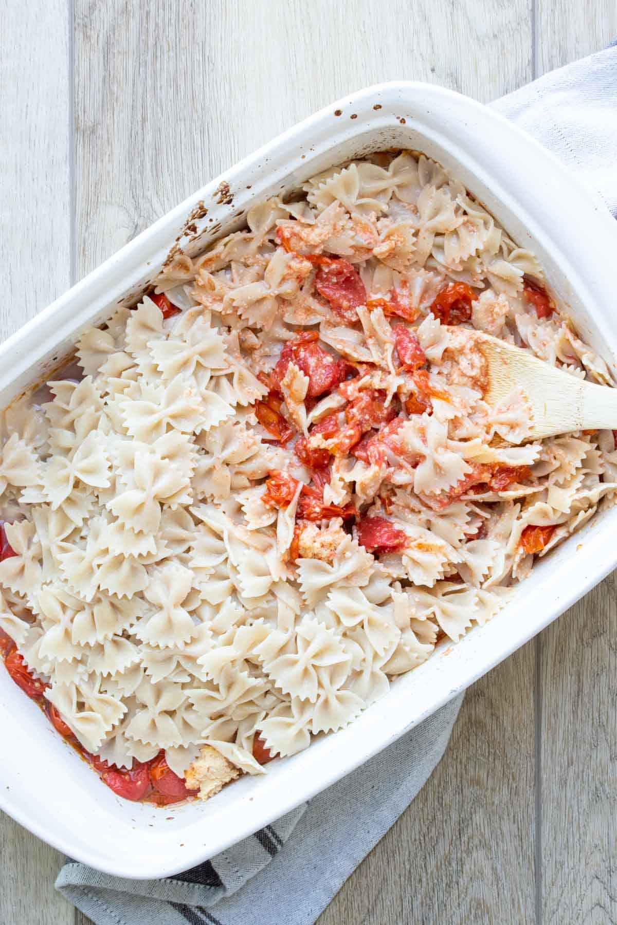 White baking dish with a wooden spoon mixing bowtie pasta in a feta tomato mix