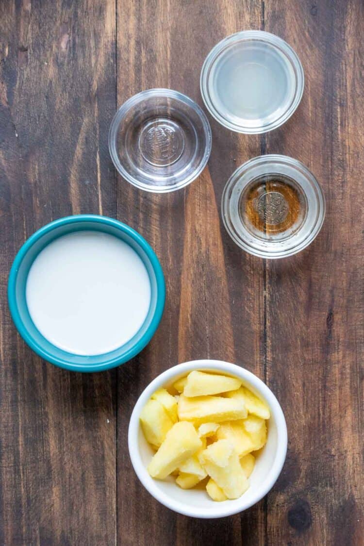 Different colored bowls with ingredients inside for making a pina colada smoothie on a wooden table