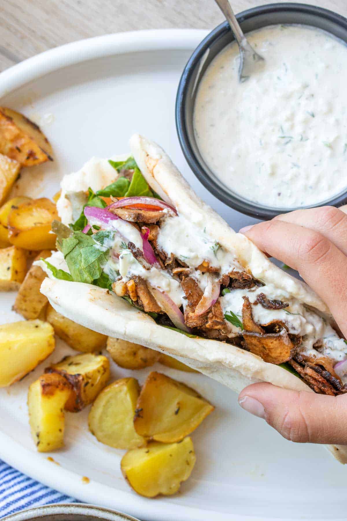 Hand holding a pita sandwich stuffed with sliced portobello, lettuce, tomato, onion and sauce.