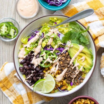 A white bowl with a taco salad drizzled with light pink sauce sitting on a yellow checked towel