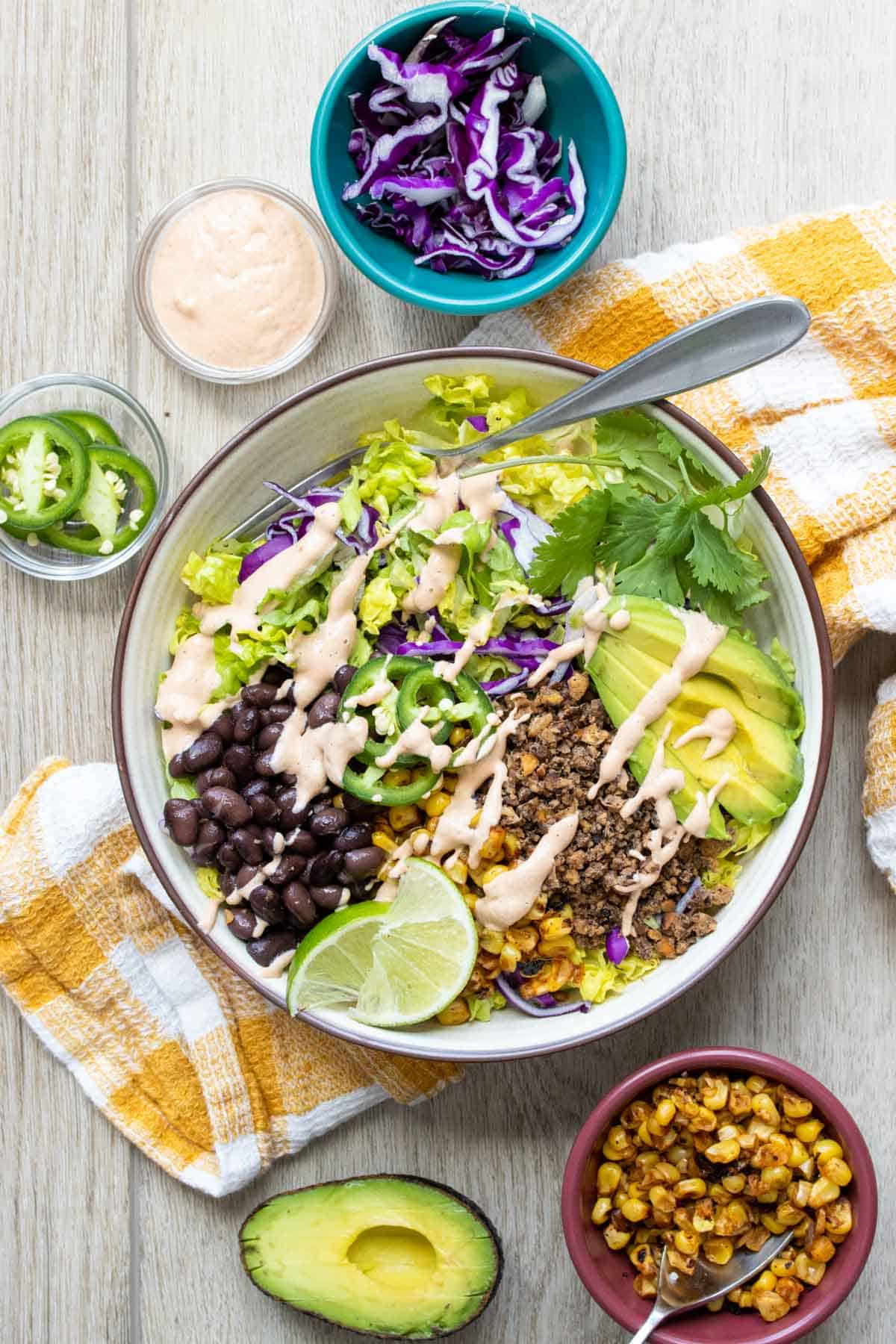 A white bowl with a taco salad drizzled with light pink sauce sitting on a yellow checked towel