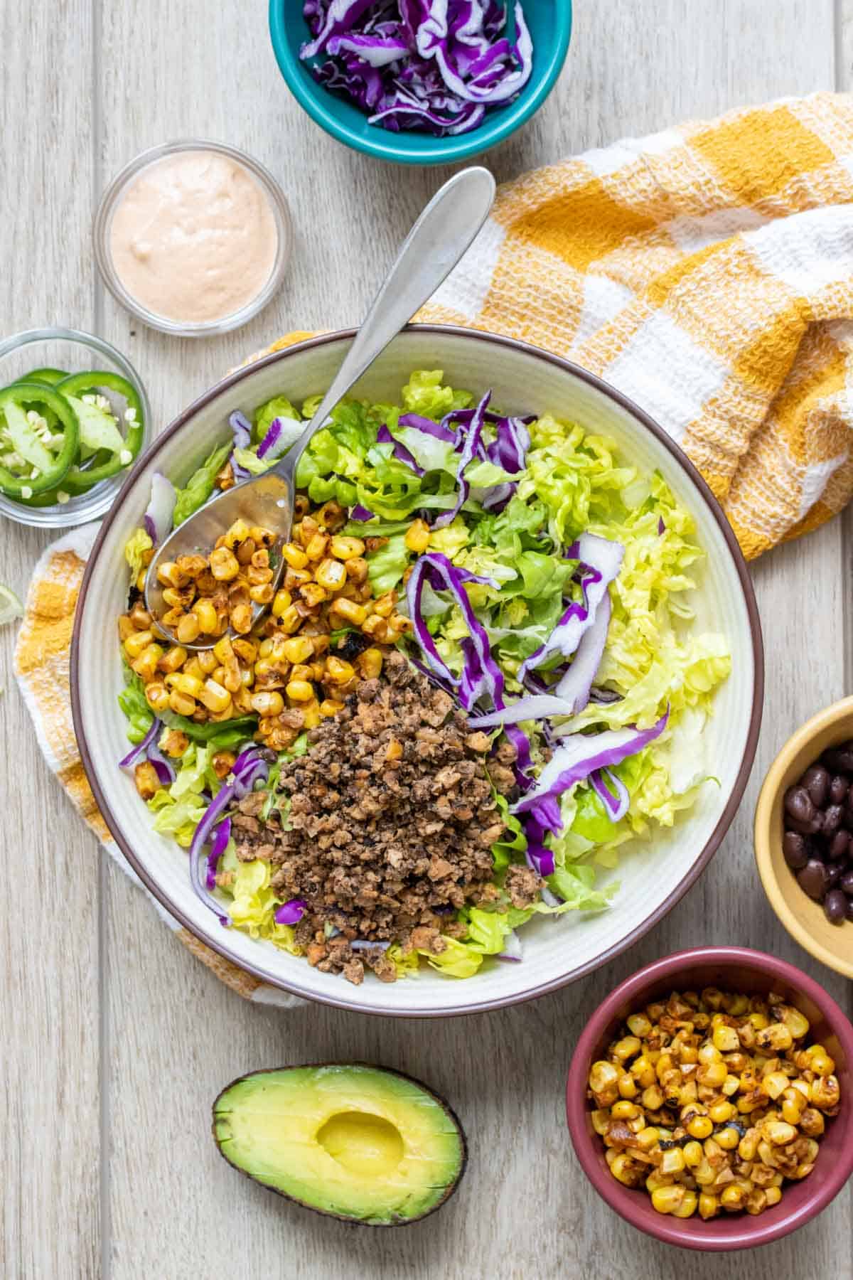 White bowl with lettuce, red cabbage, taco meat and corn on a wooden surface next to toppings