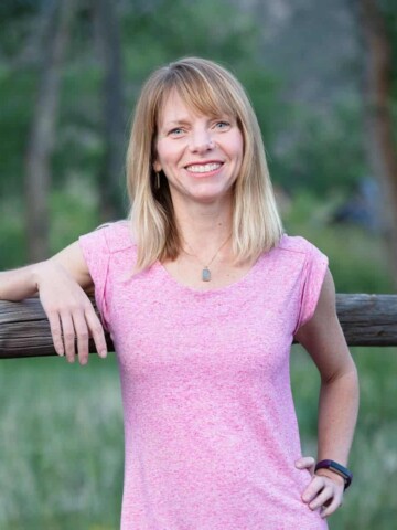 Woman in a pink shirt smiling and leaning on a piece of horizontal wood outside