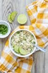 Cilantro lime rice in a white bowl on a yellow checkered towel next to cilantro a half a lime on a wooden surface.