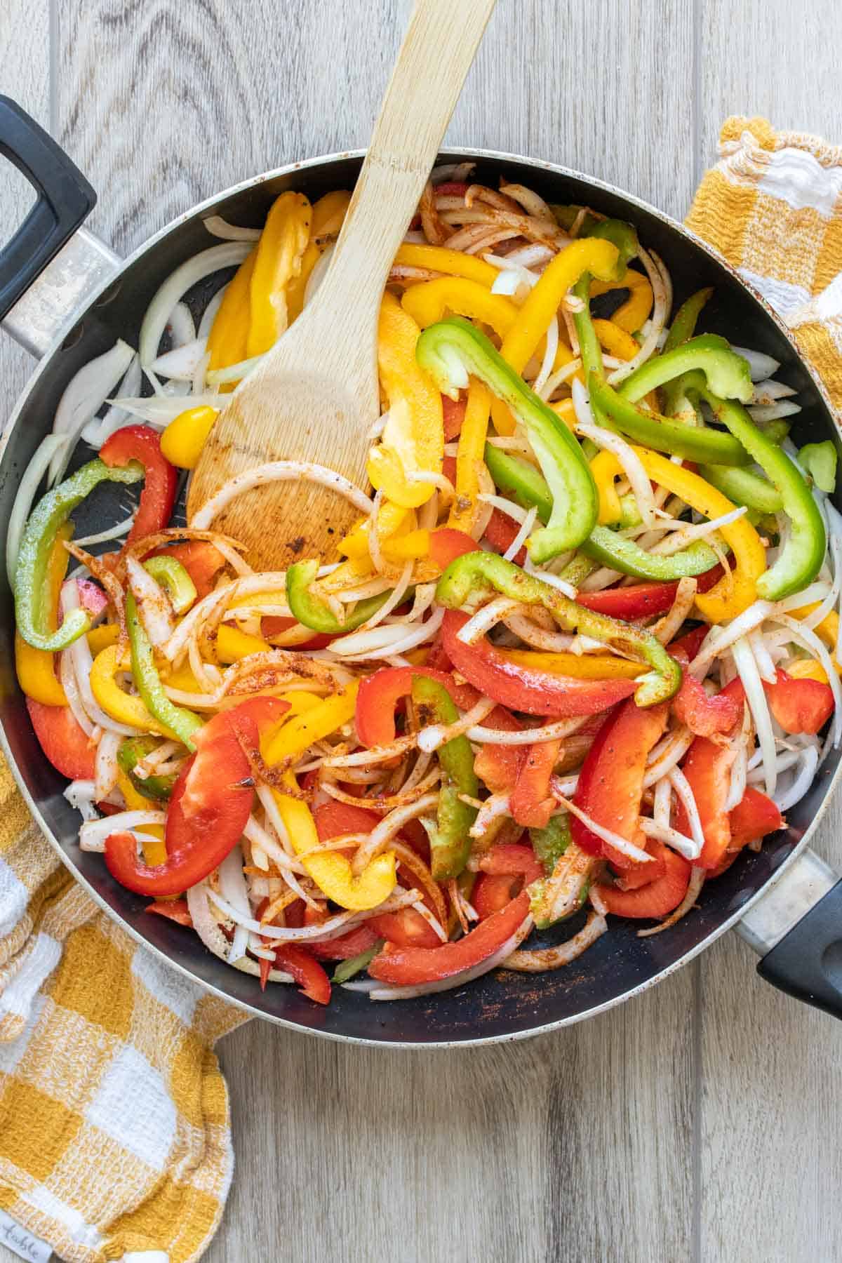 Pan with sliced peppers and onions being mixed with a wooden spoon.