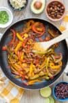 Red, yellow and green peppers being sauteed in a pan with a wooden spoon on a yellow checkered towel.