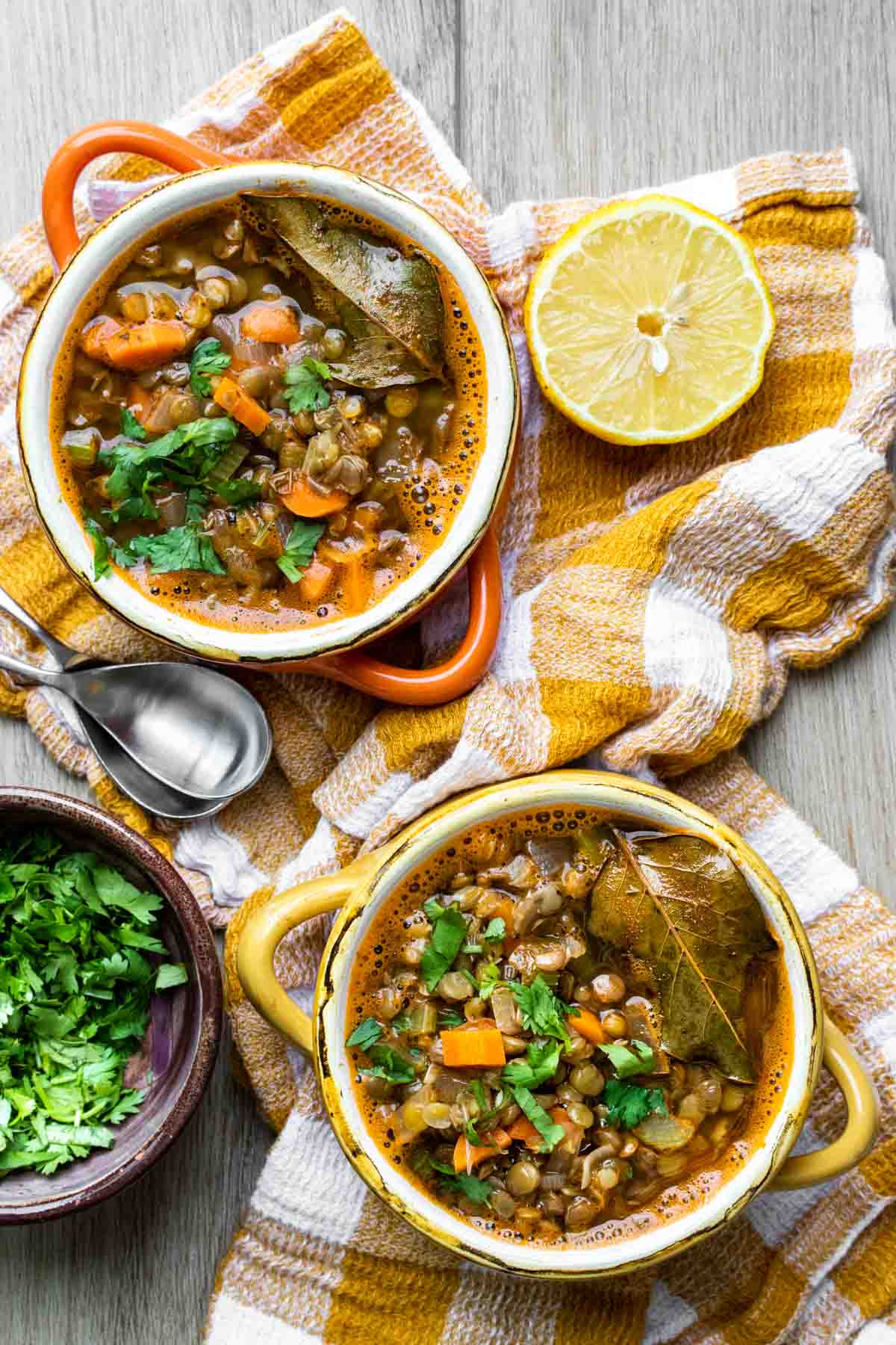 An orange and a yellow soup bowl filled with lentil veggie soup on a yellow checkered towel next to toppings