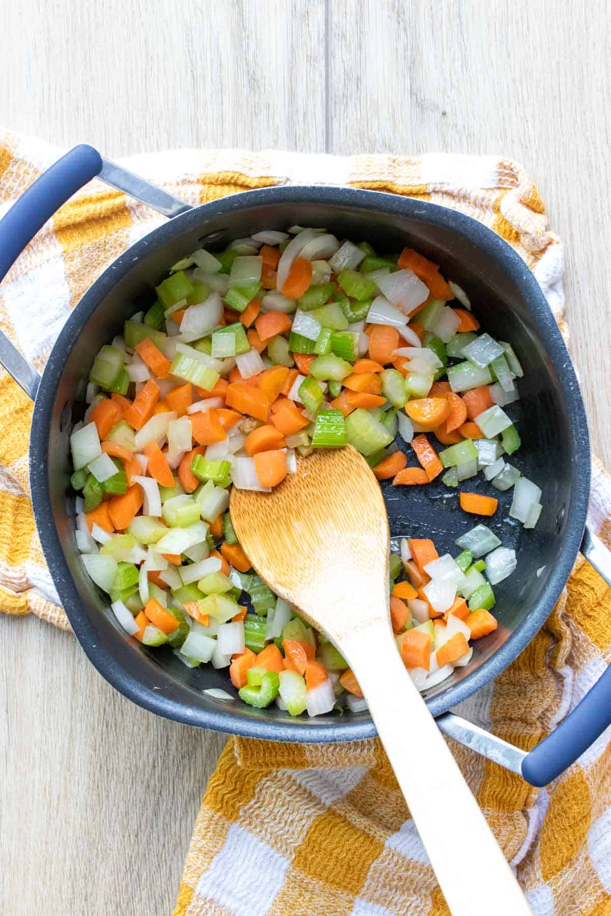 Wooden spoon mixing carrots, celery and onion in a pot on a checkered towel