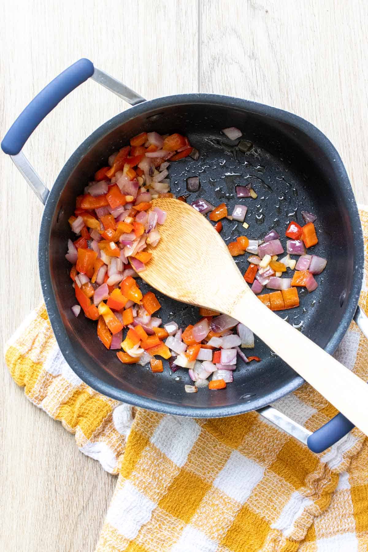 Chopped red peppers and onions being sauteed in a black pot and mixed with a wooden spoon
