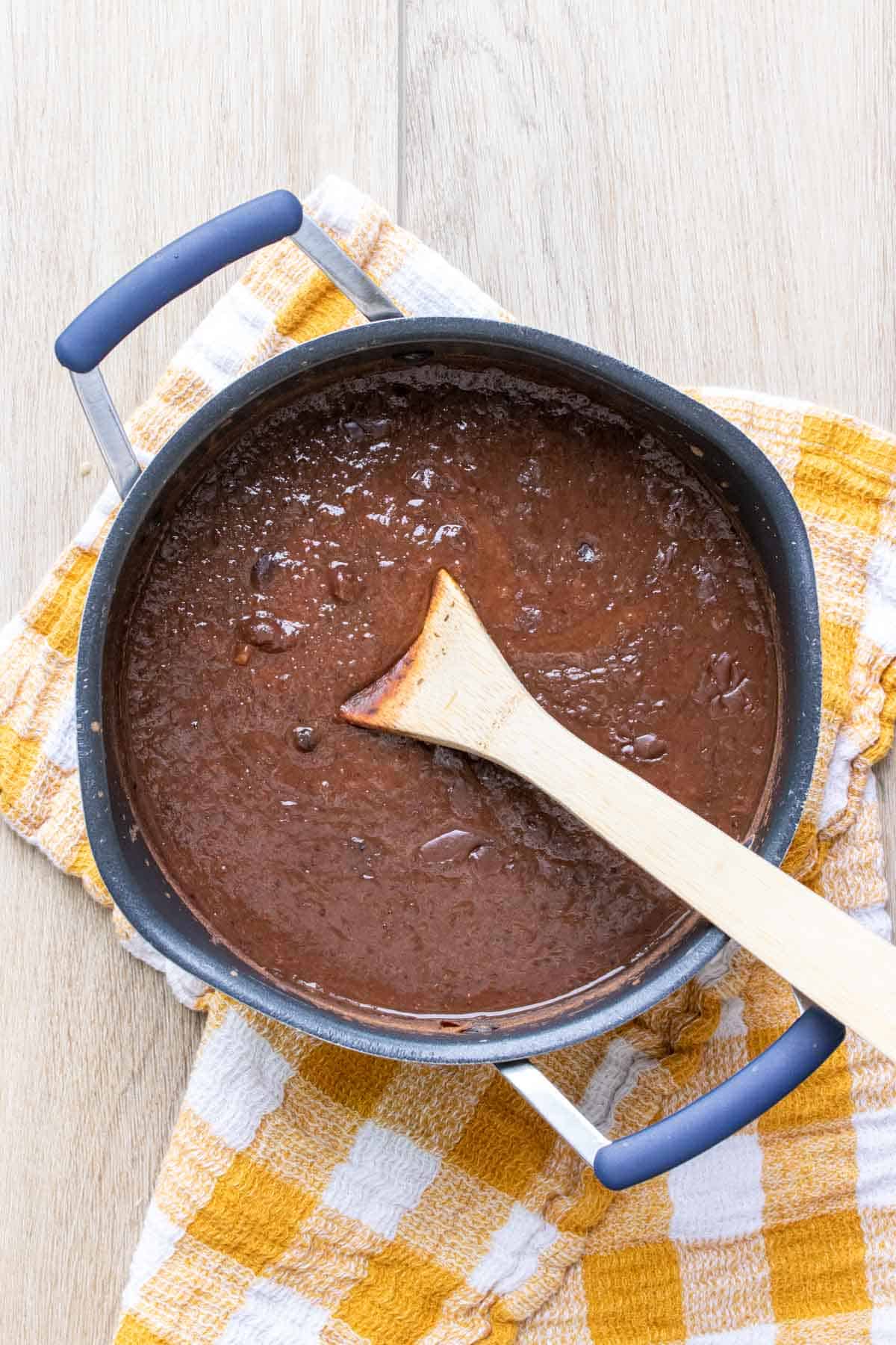 Wooden spoon mixing a pureed black bean soup in a pot on a checkered towel