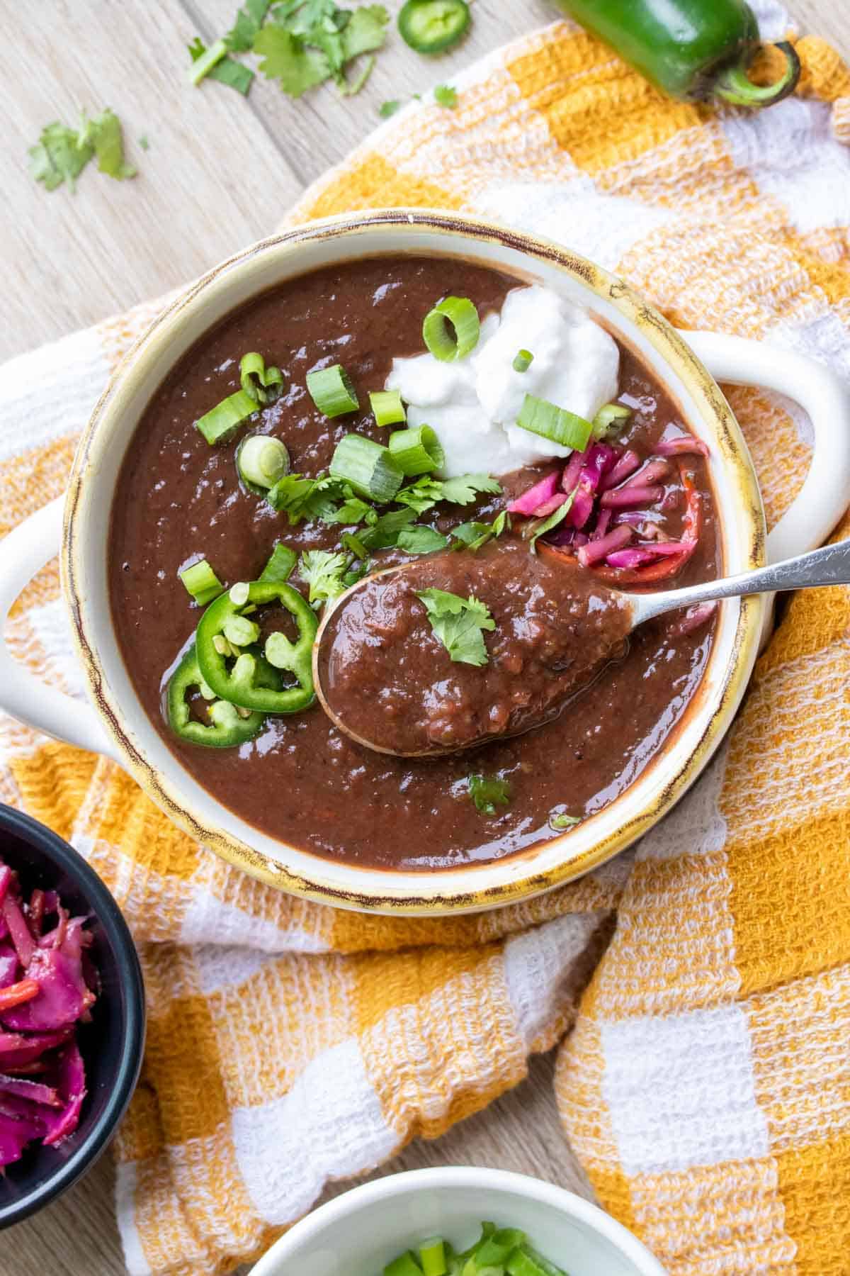 A spoon getting a bite of black bean soup from a white soup bowl sitting on a yellow and white checkered towel