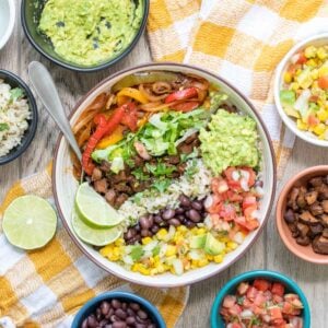 A fully loaded burrito bowl sitting on a yellow checkered towel surrounded by ingredients.