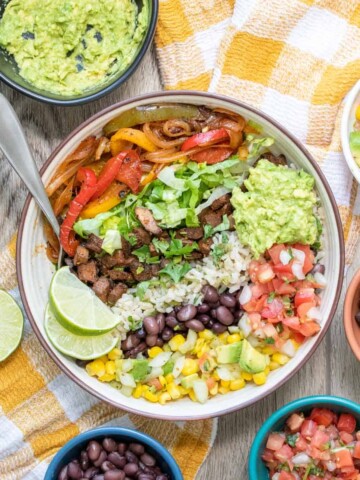 A fully loaded burrito bowl sitting on a yellow checkered towel surrounded by ingredients.