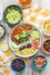 A Chipotle like burrito bowl on a yellow checkered towel surrounded by ingredients.