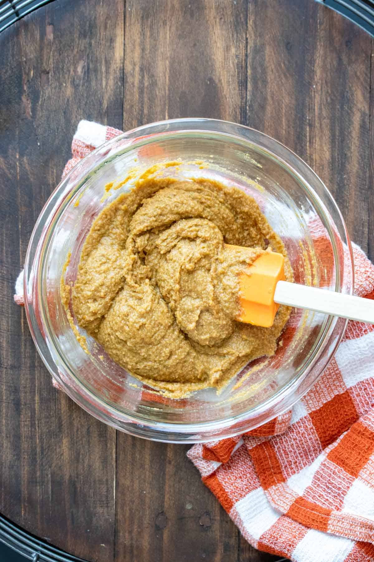 Spatula mixing pumpkin cookie batter in a glass bowl on top of an orange checkered towel