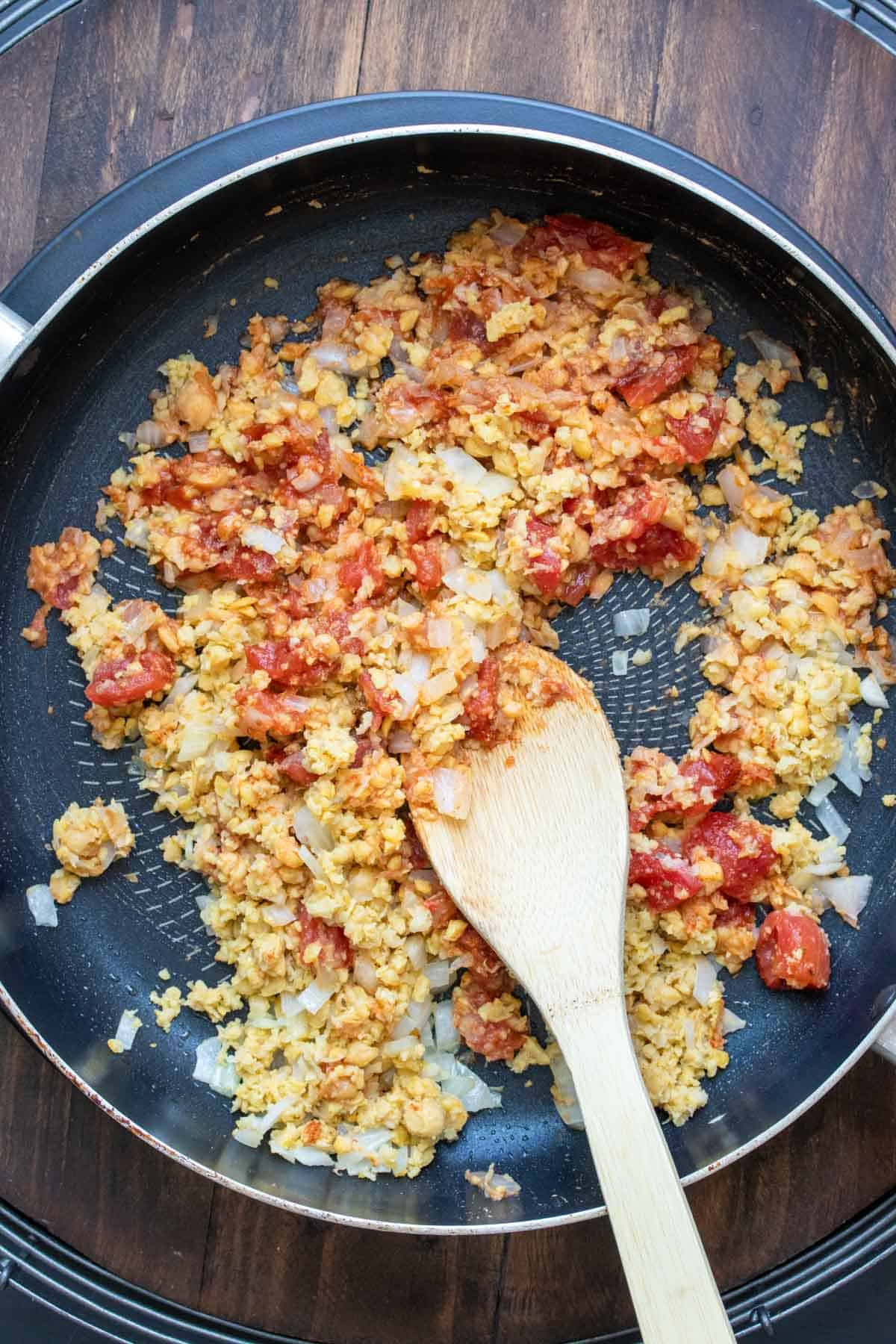 Wooden spoon mixing chopped chickpeas and chopped tomatoes in a pan