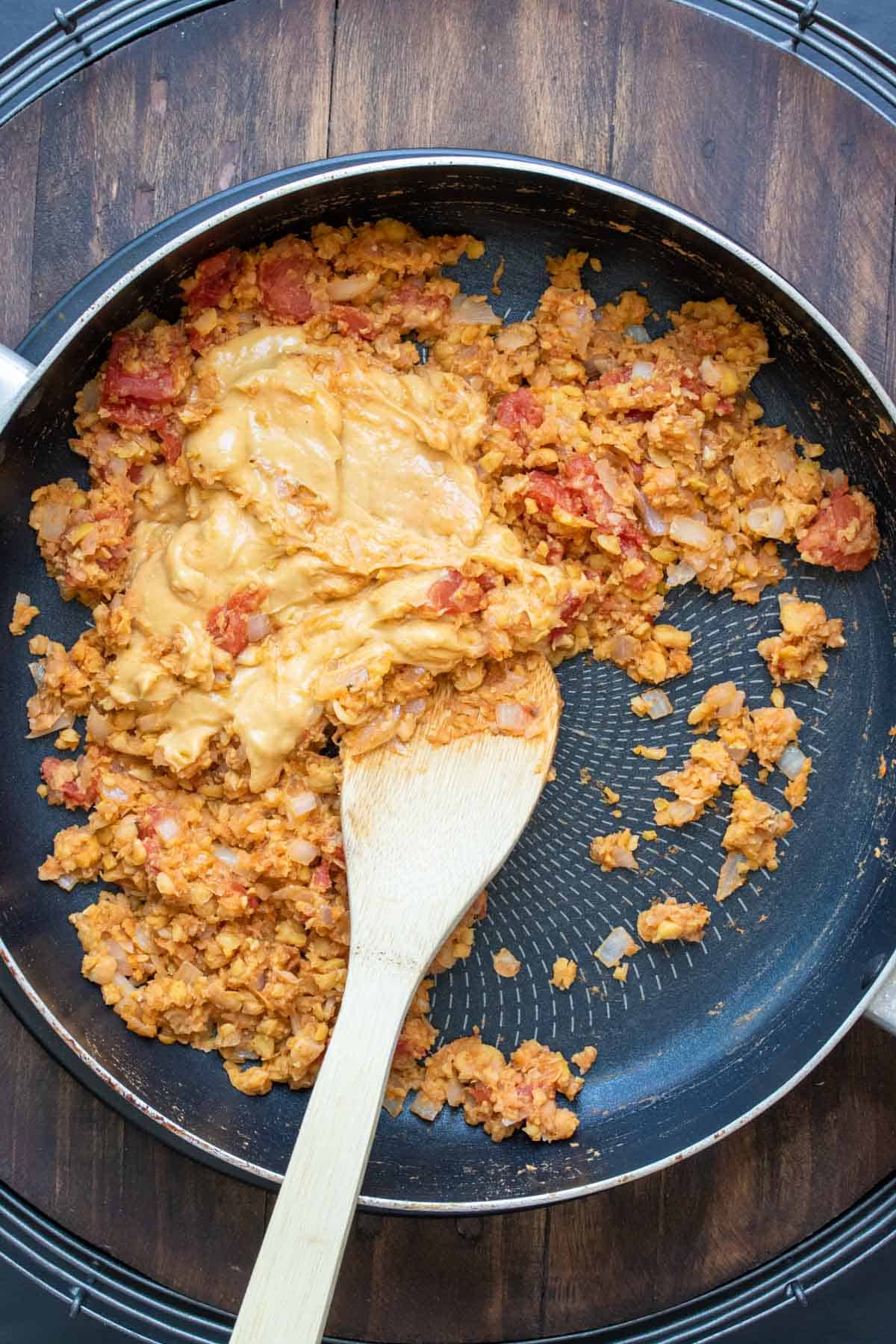 Wooden spoon mixing cheesy sauce into chopped chickpeas and chopped tomatoes in a pan