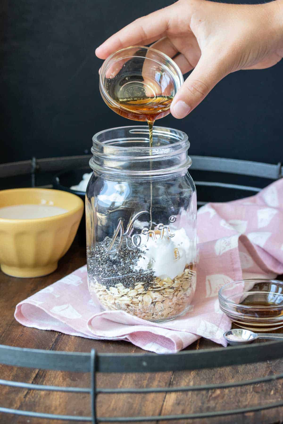 Hand pouring maple syrup in a glass jar filled with oats and chia seeds