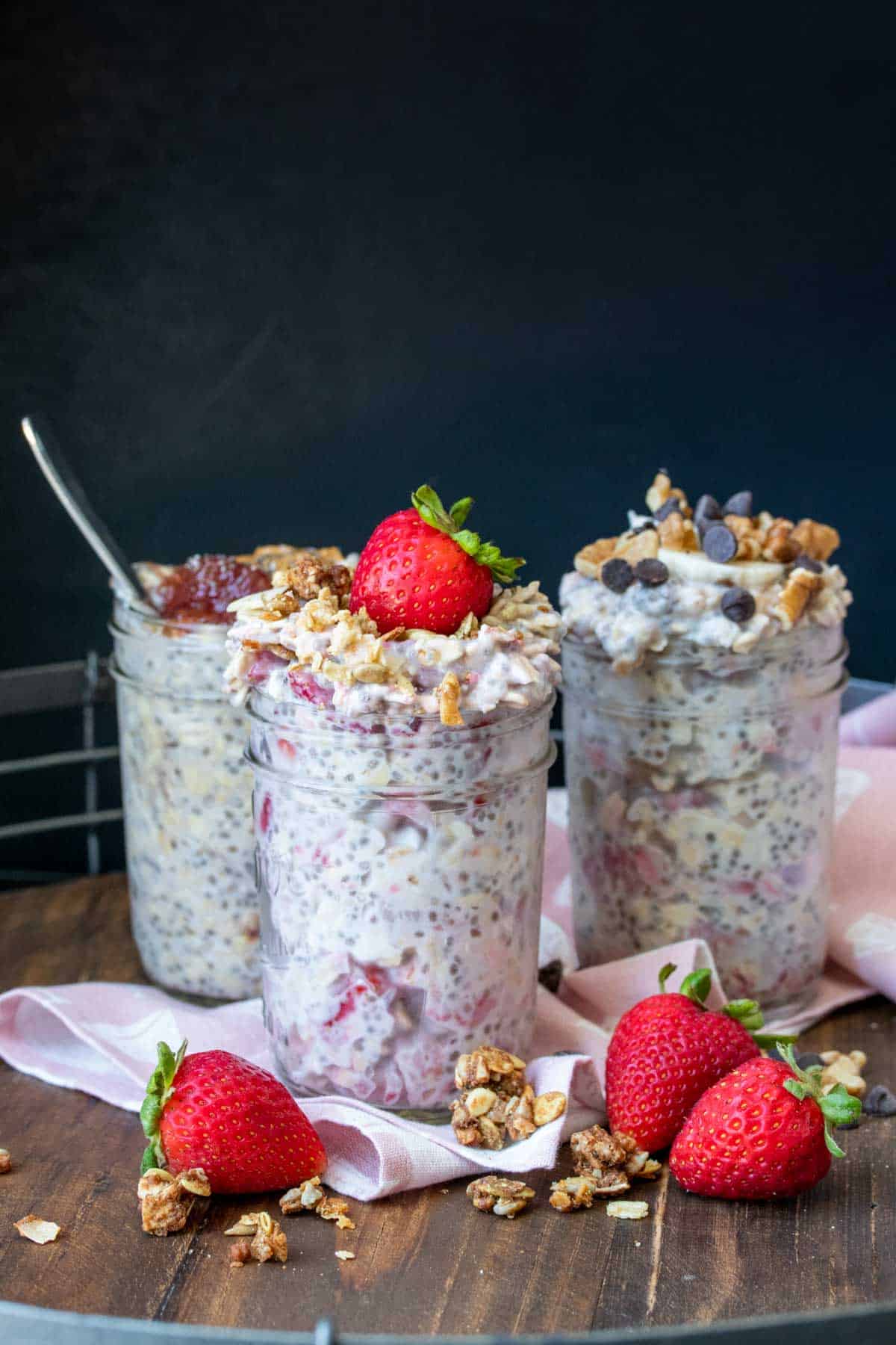 Three glass jars filled with different flavors of overnight oats sitting on a pink napkin on a wooden surface