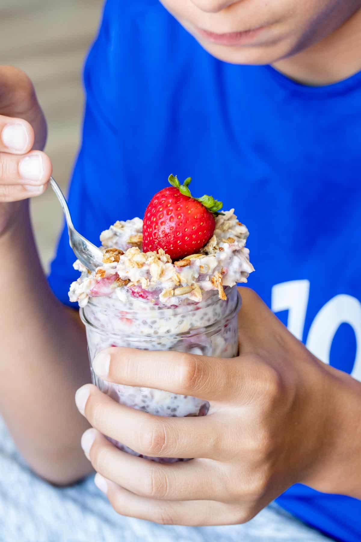 Close up of a boy getting a bite from a jar of overnight oats topped with fresh strawberries