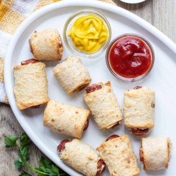 A white plate with ketchup and mustard in dipper jars and pigs in a blanket