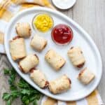 Pig in a blanket sitting on a white plate with dips or ketchup and mustard.