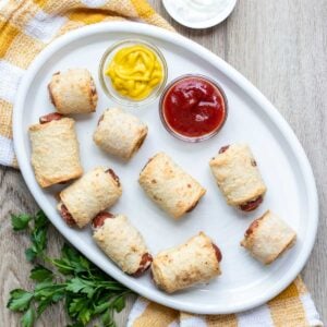 Pig in a blanket sitting on a white plate with dips or ketchup and mustard.