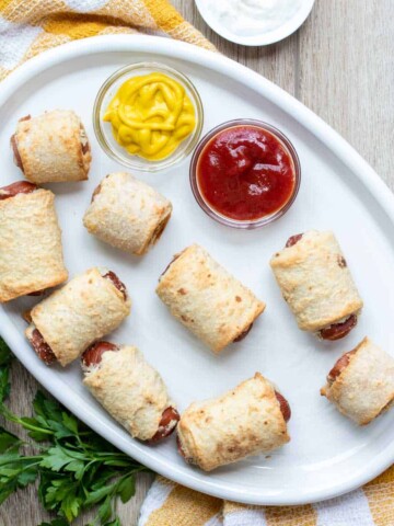 Pig in a blanket sitting on a white plate with dips or ketchup and mustard.