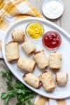 Pigs in blankets finger food on a white platter with ketchup and mustard in small glass bowls on a checkered towel.