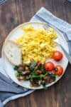 Scrambled eggs on a plate next to sauteed veggies, fresh tomatoes and bread.