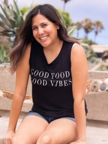 A woman with long brown hair and black tank top smiling at the camera sitting on a bench