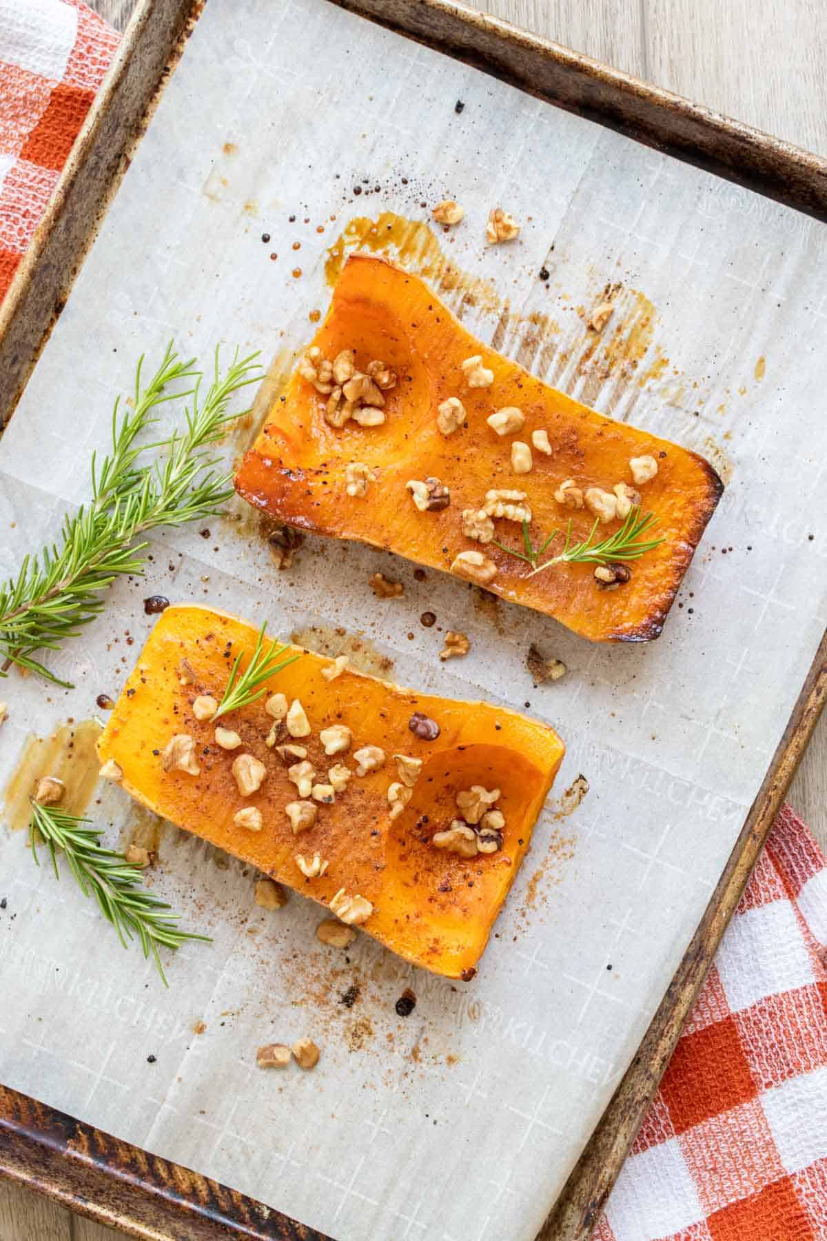 Two halves of a butternut squash on a baking sheet with nuts and rosemary.