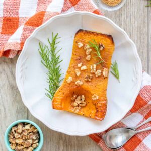 White plate with a baked half of butternut squash covered in nuts and rosemary sprigs.